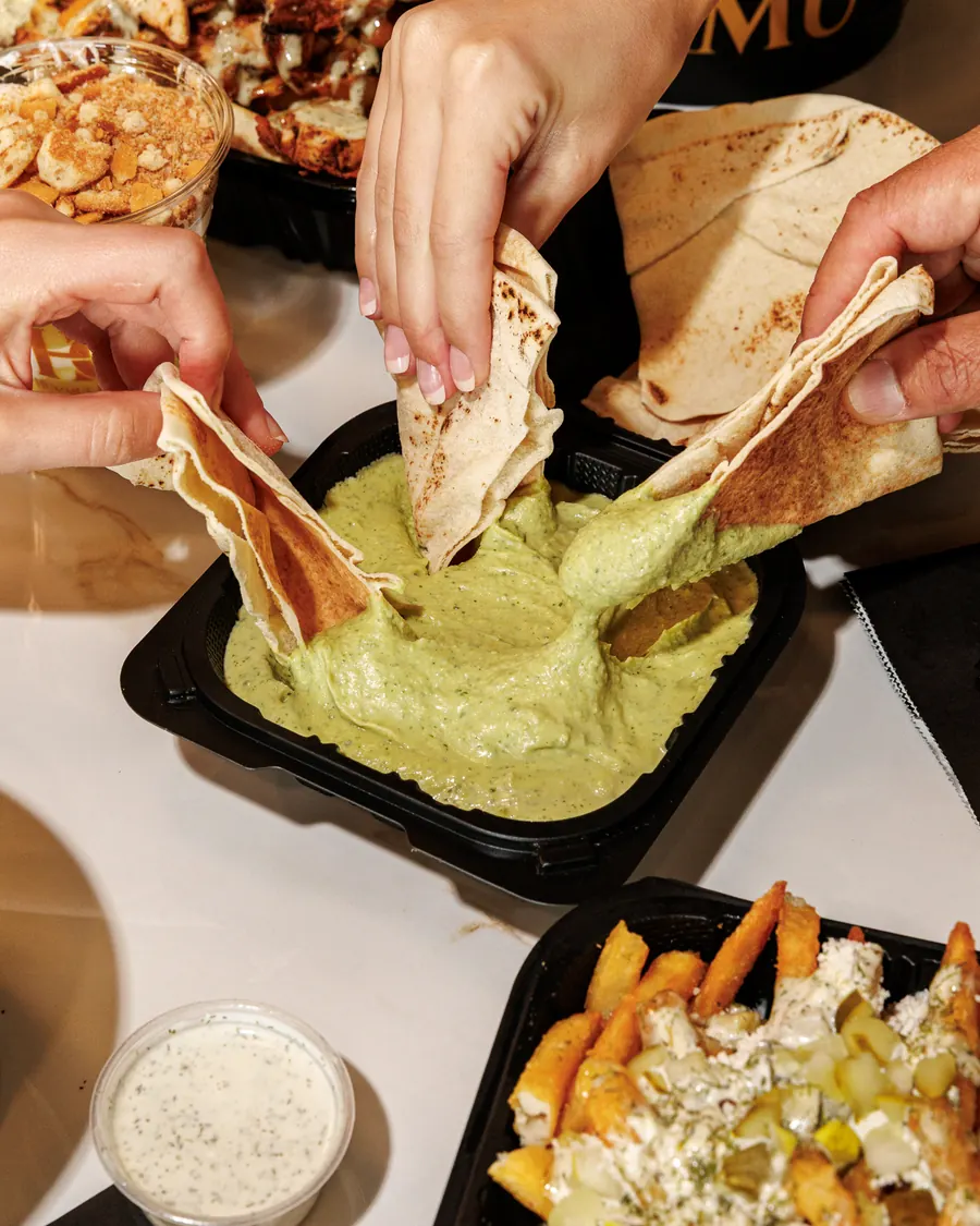 Hands dipping pita bread into a tray of cilantro jalapeño hummus beside dill fries